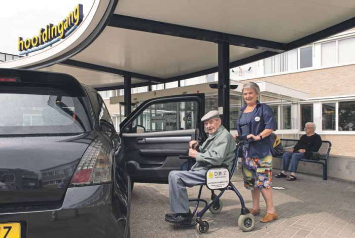 Vrijwilliger helpt ouder persoon in rolstoel naar de auto voor de hoofdingang van het ziekenhuis.