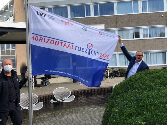 Bestuurder Ton Hazekamp hijst met hulp van beveiliger Heico Harmsen de vlag Horizontaal Toezicht in de mast van het Slingeland Ziekenhuis.